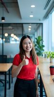 Happy Asian businesswoman giving thumbs up gesture in modern office workspace with glass walls and natural lighting for corporate success concepts.