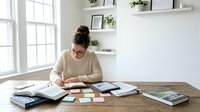 Young woman working at home office desk with books and documents, studying or planning in bright modern workspace with floating shelves and natural light.