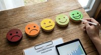 Colorful emoji feedback faces from sad to happy on wooden desk with customer satisfaction survey and tablet showing business analytics charts.
