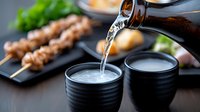 Pouring sake into traditional black ceramic cups with grilled yakitori skewers in background, creating authentic Japanese dining atmosphere and culinary experience.