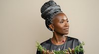 Mature African woman with gray braided hair wearing black shirt and green plants, studio portrait against neutral background.