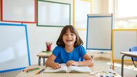 Happy young girl student smiling at desk in bright classroom with colorful whiteboards and school supplies, ready for learning and education activities.