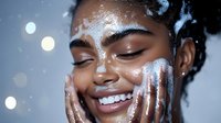 Young African American woman with glowing skin and bright smile applying white cream moisturizer on face against blurred bokeh lights background.