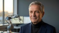 Mature male professional with gray hair and light complexion wearing dark blazer and black turtleneck, seated indoors with modern workspace and equipment visible in soft natural light.
