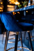 Elegant navy blue velvet bar stool with quilted pattern and gold accents against blurred bokeh lights, capturing luxury restaurant or lounge atmosphere.