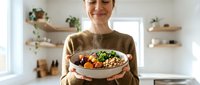 Woman in brown sweater holding bowl of roasted vegetables and grains in modern kitchen with floating shelves and plants, healthy meal preparation and home cooking.