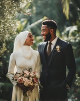 Happy Muslim bride and groom laughing together outdoors on wedding day with bouquet and formal attire in natural garden setting.