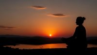 Woman silhouette meditating at sunset by lake with orange sky and clouds. Peaceful yoga practice during golden hour for wellness and mindfulness concepts.