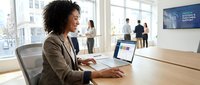 Professional African American businesswoman working on laptop in modern office conference room with colleagues in background during meeting.