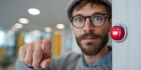 Bearded man with glasses and beanie pressing emergency button with red light in modern office hallway, focus on finger touching alarm system.