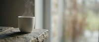 Steam rising from white ceramic mug on rustic stone surface near window with blurred outdoor background, suitable for beverage and lifestyle designs.