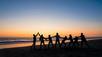 Silhouetted group of diverse people holding hands in a line on beach at sunset, celebrating unity and teamwork together by ocean waves.