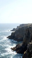 Dramatic rocky coastal cliffs rise above turquoise ocean waters with crashing waves below. Rugged basalt formations create striking vertical columns along the shoreline, ideal for travel and nature projects.