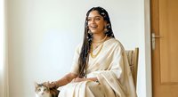 Young woman with South Asian ethnicity, long braided hair with beads, wearing gold-toned satin robe, holding tabby cat while seated indoors. Lifestyle portrait for fashion, beauty, pet care, and home decor content.