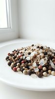 Mixed peppercorns and sea salt crystals on white plate in bright kitchen setting for culinary seasoning and cooking applications.