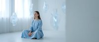 Young woman in blue robe meditating in minimalist white room with floating geometric shapes creating peaceful spiritual atmosphere for wellness content.