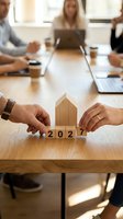 Hands holding wooden house model with 2027 blocks during business meeting, representing real estate planning and future property investment strategy.