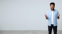 Man with dark hair and beard, appearing to be of Middle Eastern or South Asian descent, wearing a light blue button-up shirt over a white t-shirt and dark pants, gesturing expressively with both hands while looking at camera against neutral background.