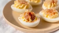 Creamy deviled eggs topped with crispy bacon bits and paprika served on ceramic plate, close up view of classic appetizer for parties and gatherings.