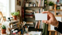 Close-up of hand holding blank white business card in modern home office with shelves, plants, and desk supplies visible in background.