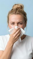 Young woman with blonde hair in top bun, Caucasian ethnicity, blowing nose with tissue against blue background for cold, flu, allergy or illness concepts.