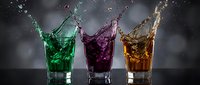 Three colorful cocktail glasses with dynamic liquid splashes in green, purple and amber against dark background for beverage advertising and bar concepts.