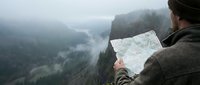 Hiker holding map while standing on mountain cliff overlooking misty valley and forested landscape below during outdoor adventure and navigation.