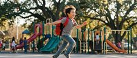 Young Asian boy in striped shirt and red backpack running joyfully at colorful playground during golden hour with trees and play equipment in background.