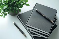Stack of black hardcover notebooks with pens and green plant on white desk for business planning and office organization.
