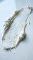 Creamy white skincare product smear with textured strokes and droplets on light background, suitable for cosmetics and beauty packaging design.