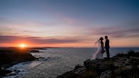 Bride and groom standing on rocky coastal cliff at sunset, silhouetted against golden sky and ocean horizon. Wedding photography.