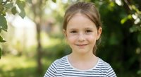 Young Caucasian girl with brown hair in striped shirt smiling outdoors in natural park setting with green foliage background for family lifestyle content.