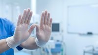 Healthcare professional wearing protective gloves in medical facility showing stop gesture for safety protocols and infection control measures.
