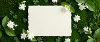 Blank white card surrounded by delicate white flowers and green leaves on natural moss background for wedding invitations, greeting cards and spring designs.