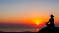 Woman silhouette meditating in lotus pose on rock at sunset over ocean. Peaceful yoga practice during golden hour with colorful sky reflection on water.
