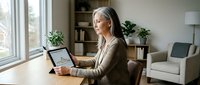 Senior businesswoman with headphones using tablet for video conference call in modern home office workspace.