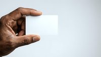Male hand with dark skin tone holding blank white card or paper against neutral background for branding and marketing mockups.
