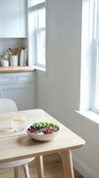 Wooden table with fresh fruit bowl and water glass in bright kitchen with natural light from window, healthy eating and nutrition concept.