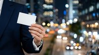 Businessman in dark suit holding blank white business card against blurred city lights at night, suitable for corporate branding and networking concepts.