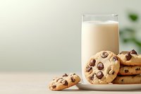 Fresh baked chocolate chip cookies stacked beside tall glass of cold milk on wooden table with soft green background, classic comfort food styling.