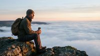 Senior man with gray hair and beard, wearing brown jacket and backpack, sitting on rocky mountain peak above clouds at sunrise, contemplating scenic landscape view.