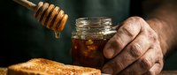 Mature man's hand holding jar of honey with wooden dipper dripping golden sweetener over fresh bread for cooking and baking.