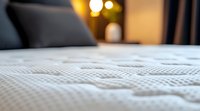 Close-up view of white textured mattress surface with soft bokeh lights in background, showing quilted pattern detail and comfort features for bedroom interior design.