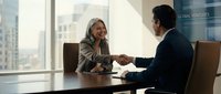 Professional businesswoman and businessman shaking hands across conference table in modern office meeting room with city skyline view through windows.