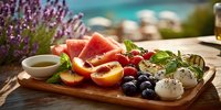 Mediterranean charcuterie board with fresh fruits, cheese, olives and herbs on wooden platter by turquoise sea for summer dining and entertaining.