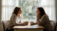 Two women sitting at table with coffee cups having conversation by bright window with sheer curtains, suitable for lifestyle, wellness, friendship and communication concepts.