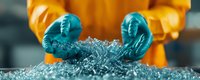 Hands in protective turquoise gloves holding recycled plastic granules against orange protective suit background, demonstrating industrial recycling process.