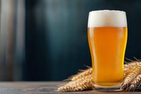 Glass of fresh draft beer with wheat ears on rustic wooden table against dark background, golden foam head and bubbles, cold refreshing beverage.