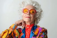 Stylish senior woman with white curly hair wearing vibrant colorful fashion outfit, orange sunglasses and bold jewelry against light background. Perfect for aging, fashion, and lifestyle content.