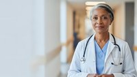 Confident mature African American female doctor in white coat with stethoscope standing in modern hospital corridor, healthcare professional portrait.